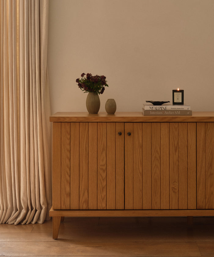 A wooden cabinet with vertical grooves stands against a cream wall. On top are dark flowers in a vase, a small ceramic vessel, books, and the Vail Glass Candle with a holiday scent. Cream curtains hang to the left.