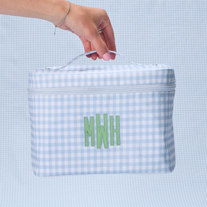 A hand holding a light blue gingham print vanity case by the top handle. The case features a custom embroidered monogram 'MWH' in green thread on the front center.