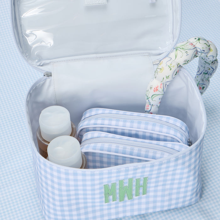An open, light blue gingham print vanity case featuring a custom embroidered 'MWH' monogram. The interior is organized with two smaller matching gingham toiletry bags and a few bottles, with a floral-patterned headband resting inside.