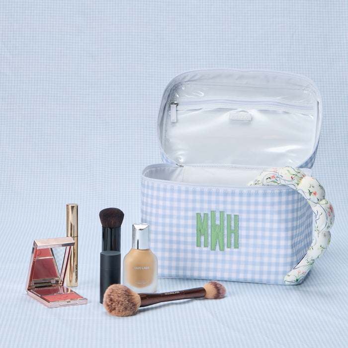 An open, light blue gingham print vanity case featuring a custom embroidered 'MWH' monogram. The interior is organized with two smaller matching gingham toiletry bags and a few bottles, with a floral-patterned headband resting inside.