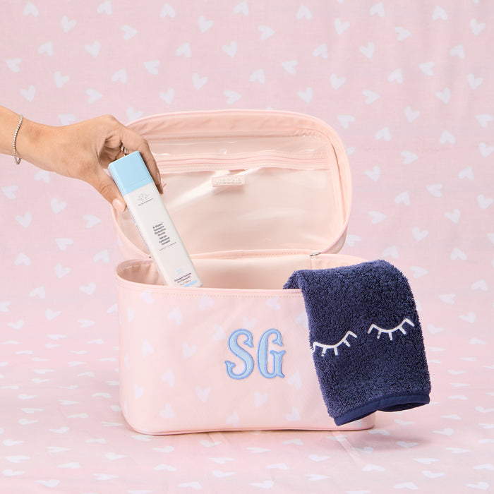 An open, light pink vanity case with a white heart pattern, featuring a blue 'SG' monogram embroidered on the front. A dark blue washcloth with an eyelash embroidery design is draped over the edge, and a hand is shown holding a Drunk Elephant serum bottle inside the case.