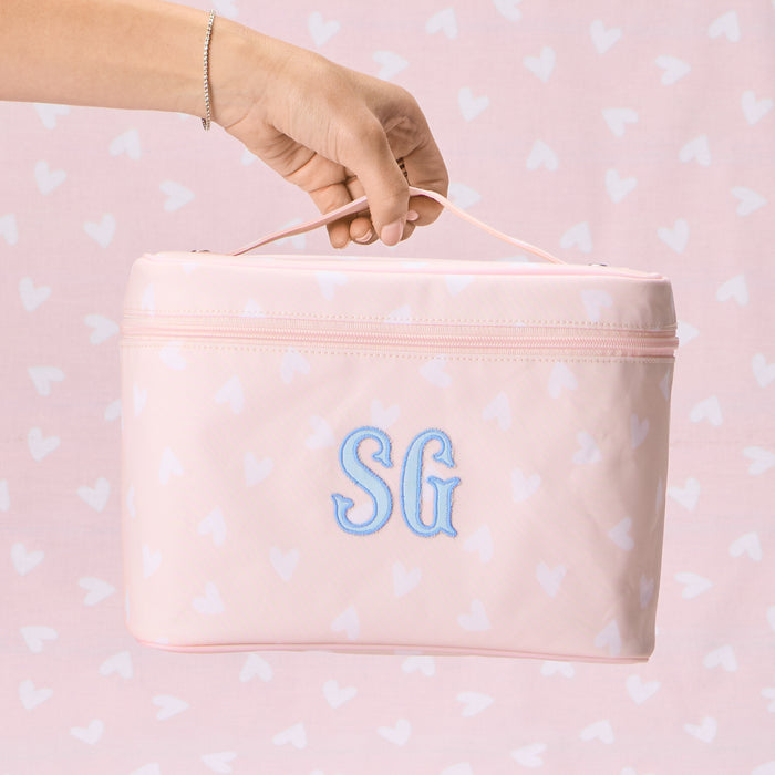 A light pink vanity case with a white heart pattern, featuring a blue 'SG' monogram embroidered on the front. A dark blue washcloth with an eyelash embroidery design is draped over the edge, and a hand is shown holding a Drunk Elephant serum bottle inside the case.