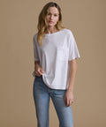 A woman modeling a white short-sleeve crewneck pocket t-shirt with light wash blue jeans, standing against a solid beige studio background.