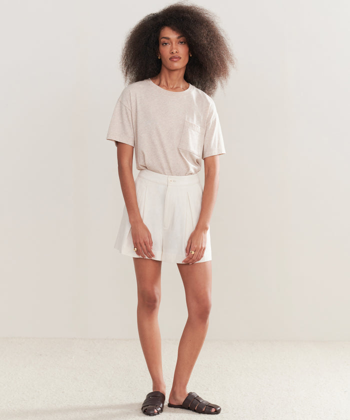 A woman with voluminous curly hair stands against a plain background, wearing the Vintage Pocket Tee in light beige, paired with white tailored shorts and dark slide sandals. Her arms rest at her sides and she wears a neutral expression.