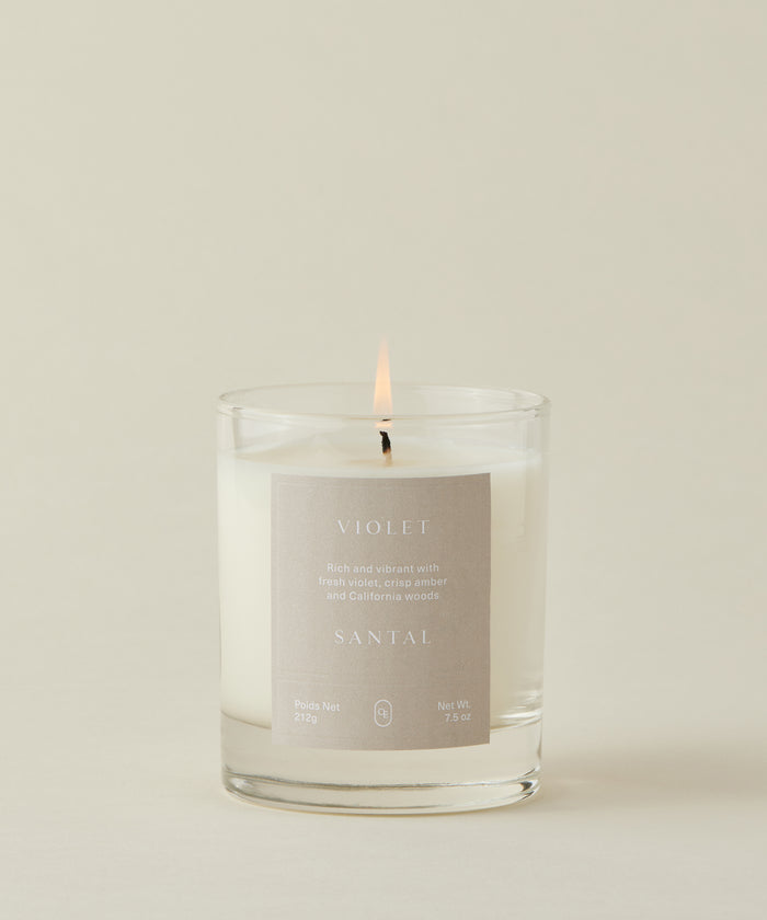 The Violet Santal Candle, in a clear glass jar with a beige label, fills the air with fresh violet, crisp amber, and California woods for coastal nostalgia against a light background.