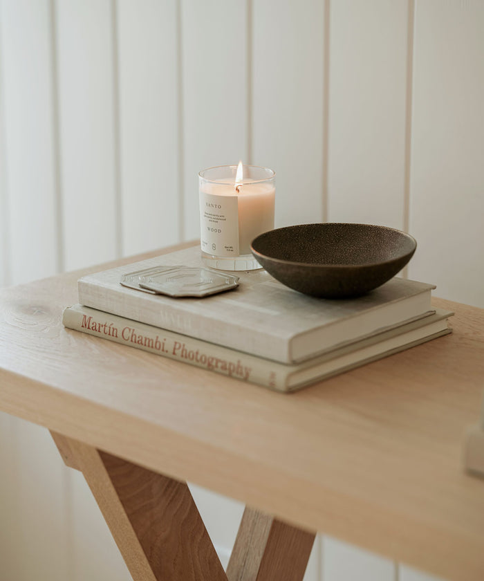 A wooden table displays stacked books, a closed notebook, a dark ceramic bowl, and the Violet Santal Candle in a glass jar—its warm glow evoking coastal nostalgia against a backdrop of white paneled walls.