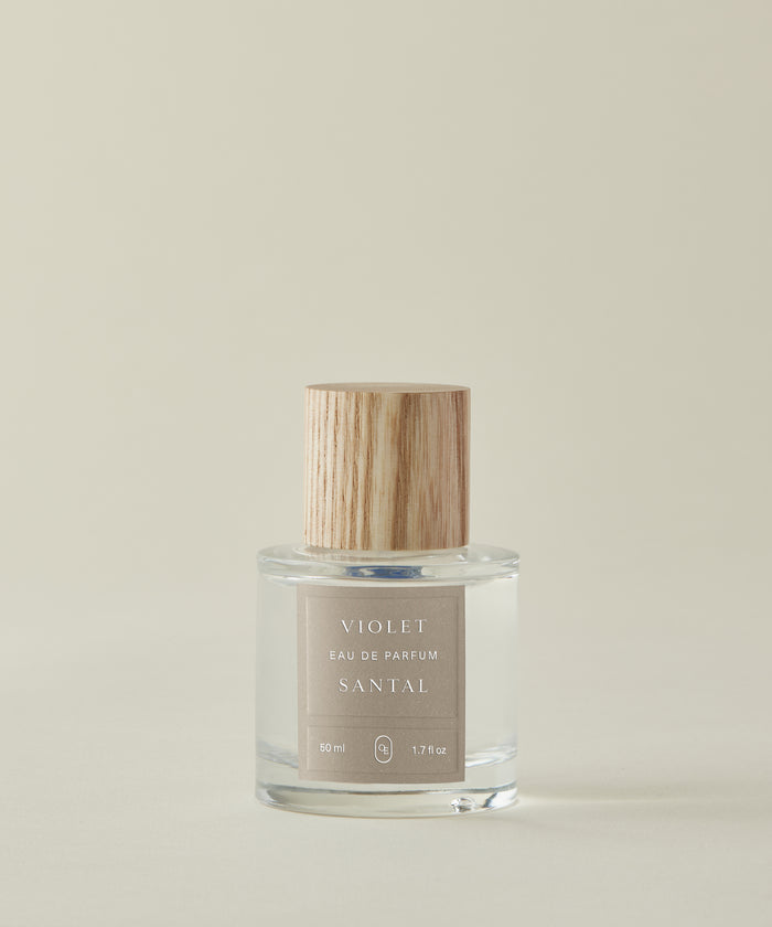 A clear glass bottle of Violet Santal Eau De Parfum with a light wooden cap is set against a plain beige background. This elegant fragrance blends woody and floral notes for a refined, clean scent.