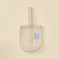 Cream canvas wine tote bag with a single strap and light blue embroidered letter M.