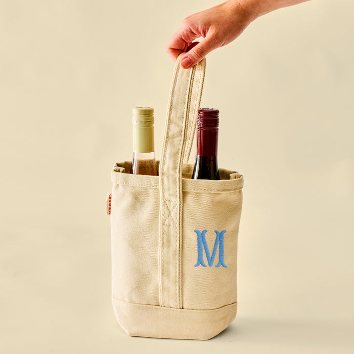 Hand holding a cream canvas two-bottle wine tote with blue M embroidery and two bottles.