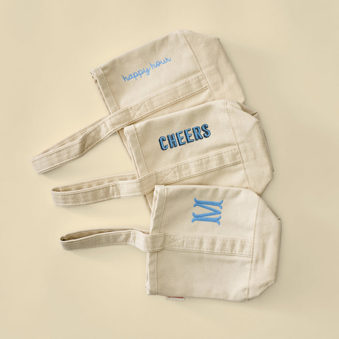 Three natural canvas wine tote bags with custom embroidery: happy hour, AWD, and a blue M.