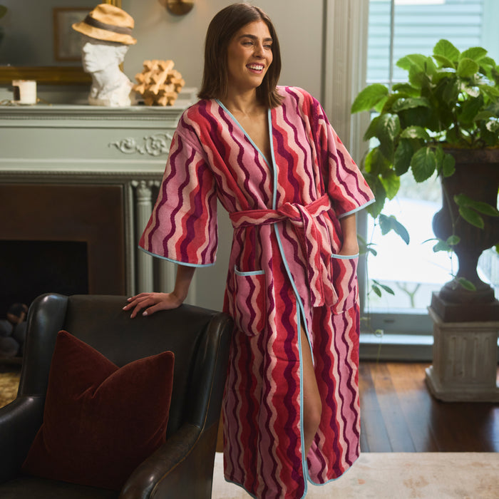 A woman wearing a pink and red wavy-patterned robe stands in a living room next to a dark leather armchair.