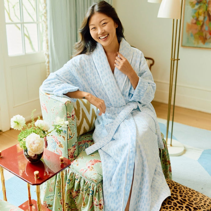 Woman in a light blue robe sitting on a floral chair in a bright room.