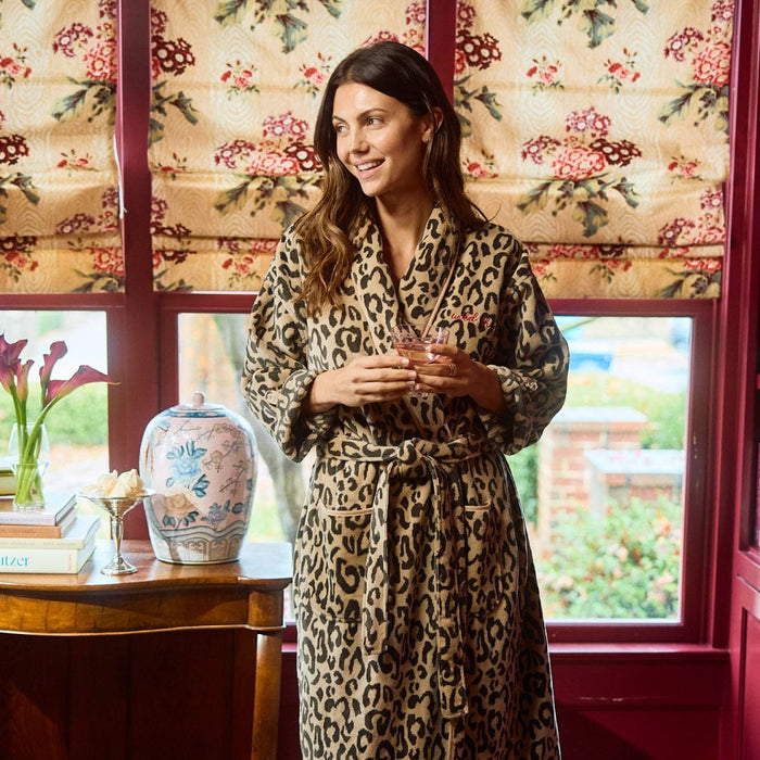 Woman in a leopard print robe holding a glass in a room with floral curtains and decor.