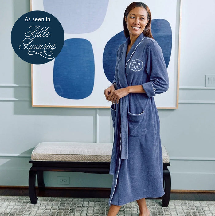 A woman wearing a blue plush bathrobe with white piping and a monogrammed 'EUC' patch on the chest, standing in a styled room with a bench and abstract wall art. A circular badge in the top left reads 'As seen in Little Luxuries'.