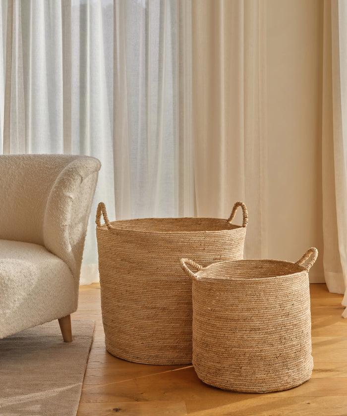 Two Woven Storage Baskets with handles rest on a wooden floor beside a cream-colored chair, with light curtains and warm natural light—perfect for adding stylish home storage.
