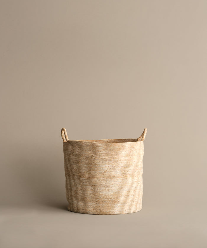 The Woven Storage Basket, made from banana leaf and featuring two handles, rests on a light brown surface with a matching background—ideal for adding style to your homes storage solutions.