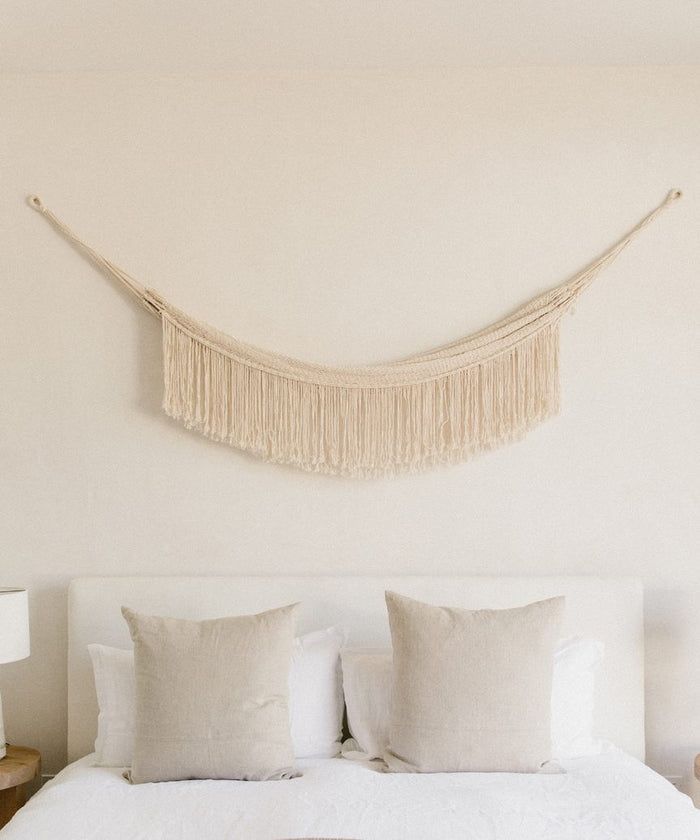 A minimal bedroom features a white headboard, two beige pillows on a bed with white bedding, and the Woven Wall Hanging in cream above the bed, adding subtle handmade decor to the plain light-colored wall.